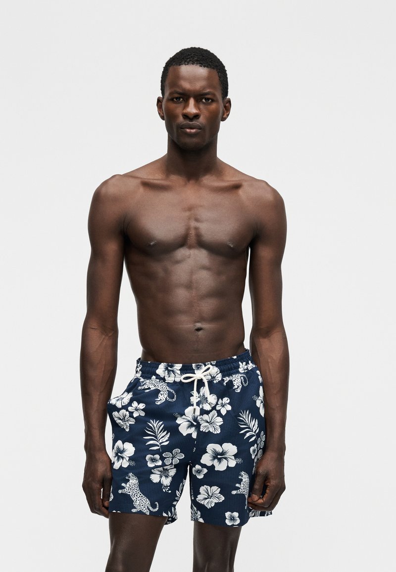 Särkimata mees, kes kannab mereväe siniseid ujumispükse valge lillelise ja leopardimustriga, seisab ühetaustalise heledalt halli tausta ees.