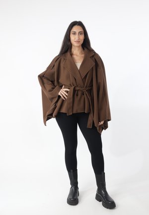 Vrouw met lang donker haar draagt een losse bruine mantel met riem, zwarte legging en zwarte enkellaarzen tegen een witte achtergrond.