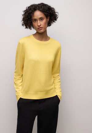 Žlutý pletený svetr s kulatým výstřihem, dlouhými rukávy a žebrovanými manžetami; doplněný černými kalhotami, které mají hladkou texturu.