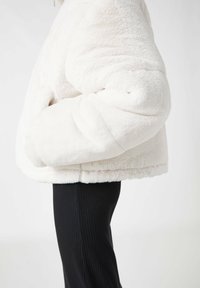 Chaqueta de pelaje sintético blanco con una superficie texturizada, ajuste holgado y forma redondeada, combinada con pantalones negros de canalé.