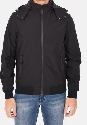 Man wearing a black zip-up jacket with a detachable hood and zippered side pockets, paired with blue jeans.