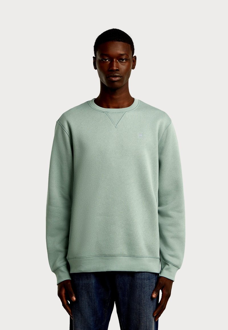 Miętowo zielona bluza z długim rękawem, okrągłym dekoltem, fakturowaną tkaniną i subtelnym logo na piersi. Połączona z dżinsami.