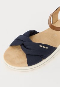Meripäivän kangasta oleva sandaali, jossa on kiertynyt yläosa, mokkapintainen pohja ja vaaleanruskea hihna. Varustettu valkoisella pohjalla ja hopeisella "Tom Tailor" -logolla.