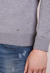 Grauer Strickpullover mit gerippten Bündchen und Saum. Mit einem metallischen "BOSS"-Logo-Akzent am Saum. Kombiniert mit ausgewaschenen blauen Jeans.