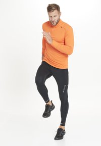 Orange langærmet træningstrøje med høj krave og sorte stræk leggings. Sorte løbesko med tekstureret overflade og snørebånd.