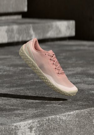 Zapatilla deportiva de malla rosa claro con suela texturizada beige suspendida sobre escalones de concreto, mostrando detalles laterales y superiores, incluyendo cordones y nombre de la marca.