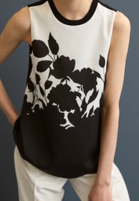 Top senza maniche con una stampa floreale nera su sfondo bianco, caratterizzato da un collo tondo nero e un tessuto morbido. Abbinato a pantaloni di colore chiaro.