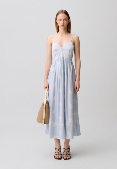 Giovane donna in piedi che indossa un vestito azzurro chiaro con spalline sottili a fiori, tiene in mano una borsa beige e indossa sandali neri con punta aperta.