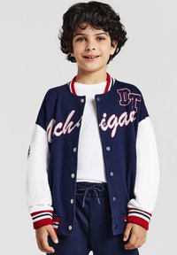 Kind mit lockigem dunklem Haar, das eine marineblaue und weiße Collegejacke mit roten gestreiften Bündchen trägt, lächelt vor einem einfarbigen Hintergrund.