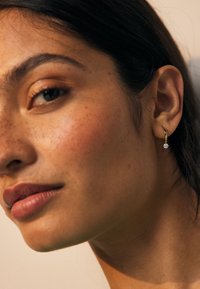 Nahaufnahme eines Frauengesichts mit Sommersprossen, natürlichem Make-up, dunklem Haar und einem goldenen Creolenohrring mit einem kleinen, hängenden Edelstein.