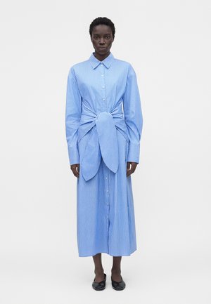 Žena nosí dlhé modro-biele pruhované šaty s gombíkmi s veľkým uzlom v páse a čiernymi topánkami, stojí.