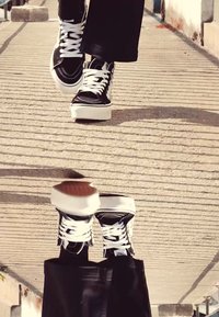Schwarz-weiße Sneakers mit dicken weißen Sohlen und weißen Schnürsenkeln, getragen mit lockeren schwarzen Hosen, auf einem strukturierten Bürgersteig gezeigt.