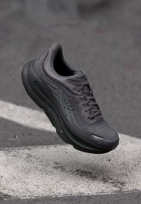 Σκούρο γκρι παπούτσι για τρέξιμο Hoka με παχιά σόλα, αιωρούμενο πάνω από άσφαλτο με λευκή οδική σήμανση από κάτω.