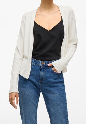 Femme portant un cardigan blanc sur un haut noir à col en V et un jean taille haute bleu, avec une main dans la poche et des bagues claires aux doigts.