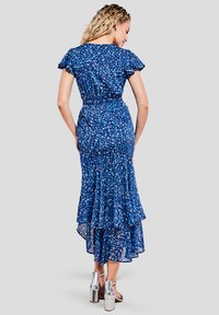 Robe florale bleue avec des manches courtes volantes, un ourlet en plusieurs niveaux, une taille cintrée et une texture transparente ; portée avec des chaussures à talons métalliques.