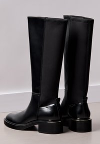 Čierne vysoké čižmy s hladkým koženým predným dielom a elastickým zadným dielom, s robustným opätkom a úchytkami pre jednoduché obúvanie.