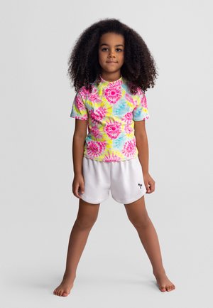 Dziecko z kręconymi włosami stoi boso, ubrane w kolorową koszulkę tie-dye i białe spodenki z małym logo palmy.