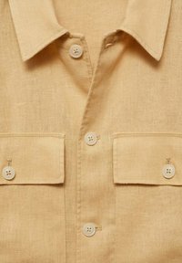Beige lněná košile s límcem, která má dvě přední kapsy a čtyři knoflíkové zapínání vyrobené z bílého plastu. Hladká textura s jemným vzorem.