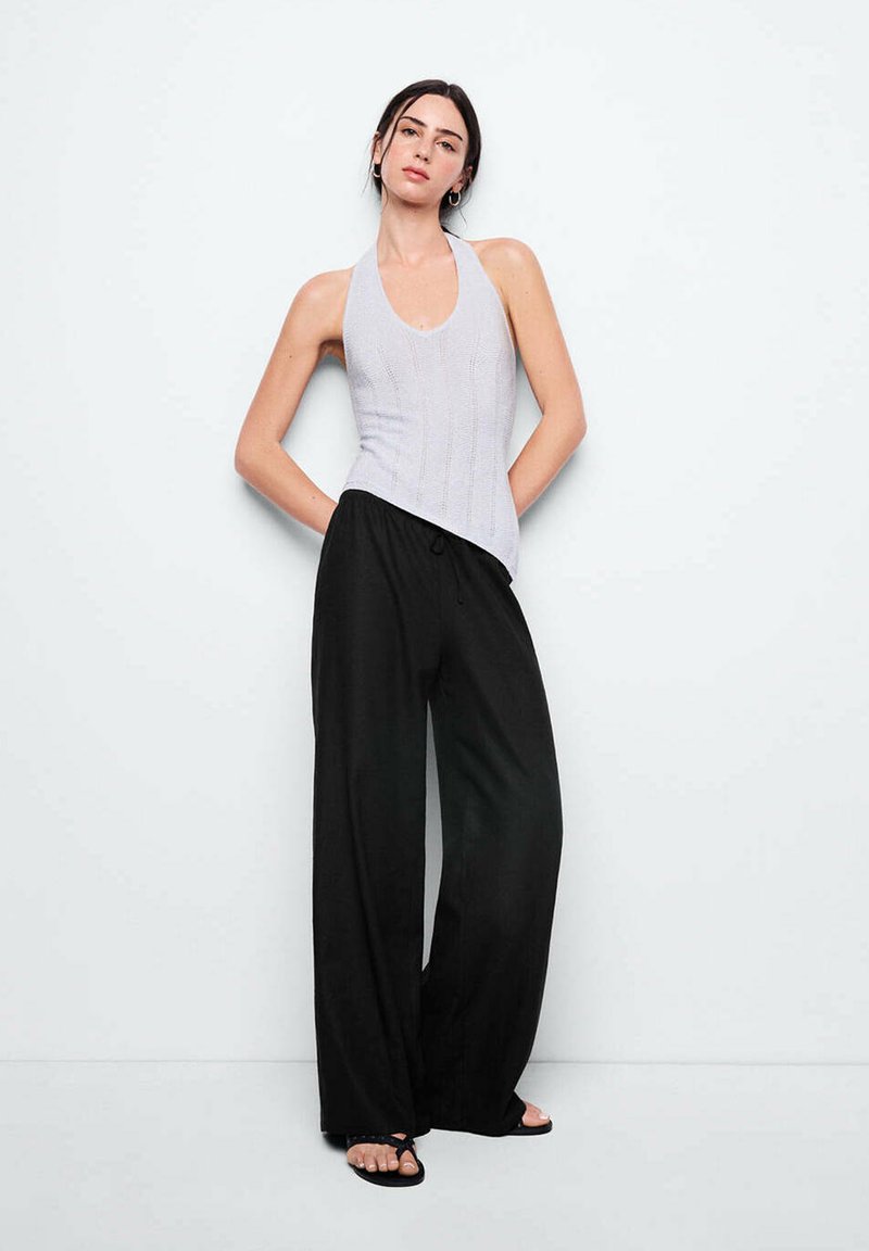 Femme debout contre un mur blanc, portant un top gris sans manches, un pantalon large noir, des sandales noires, les mains derrière le dos.