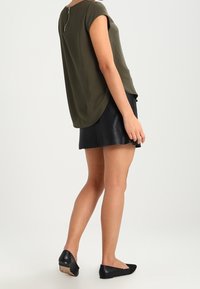Olijfgroen blouse met korte mouwen, een afgeronde zoom en een zilveren ritssluiting op de rug, gecombineerd met een zwarte leren mini rok en platte schoenen.