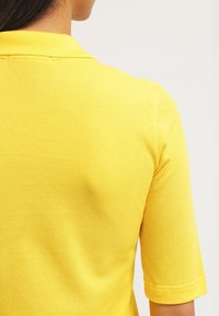 Žluté polo tričko s texturovanou tkaninou, v klasickém střihu s límcem a krátkými rukávy. Materiál vypadá jemně a lehce.