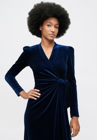 Mlad oseba z afro frizuro, oblečena v dolgo, temno modro žametno obleko z zavojo na strani, pozira z eno roko na boku.
