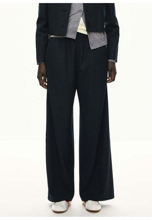 Persona lleva pantalones negros de pierna ancha, una chaqueta en capas con secciones grises y negras, y zapatillas blancas sobre un fondo blanco liso.