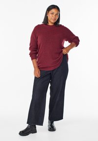 Nainen, jolla on olkapituiset hiukset, pukeutuneena burgundinväriseen neuleeseen, leveälahkeisiin tummiin farkkuihin ja mustiin nilkkabootseihin, seisoo valkoista taustaa vasten.