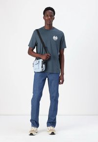 Dark teal crew-neck t-shirt with circular logo, blue denim jeans, light blue crossbody bag, and beige sneakers. Modern casual attire.