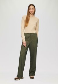 Beige langwerpige top gecombineerd met hoge taille, olijfgroene wijdvallende broeken. Schoenen zijn bruin met lichte accenten.