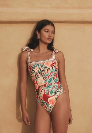 Frau mit langen dunklen Haaren trägt einen bunten Badeanzug mit gemusterten Mustern und gebundenen Schulterriemen, steht vor einer beigen Wand.