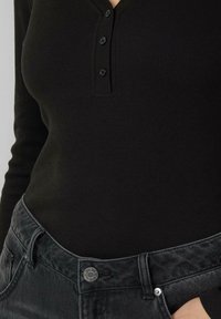 Czarna prążkowana bluzka z długim rękawem, z dekoltem typu henley zapinanym na trzy guziki, zestawiona z ciemnoszarymi spodniami dżinsowymi z widocznym srebrnym guzikiem.