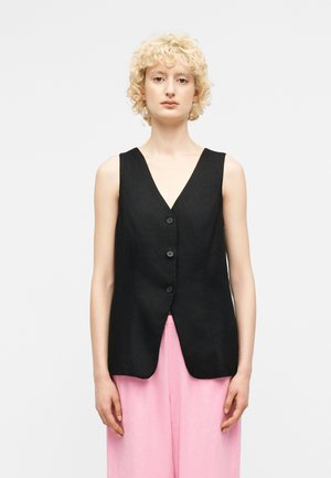 Person with short curly blonde hair wearing a sleeveless black buttoned vest and loose pink pants, standing against a white background.