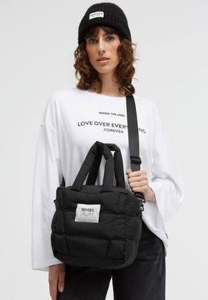 Schwarze gesteppte Handtasche mit zwei kurzen Griffen und einem abnehmbaren langen Riemen. Mit einem weißen Etikett mit der Aufschrift "SENSES". Weiche Textur.