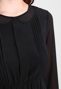 Svart klänning med plisserad design, rundad krage och genomskinliga långa ärmar. Släthand med elastiskt midjeband för en definierad silhuett.