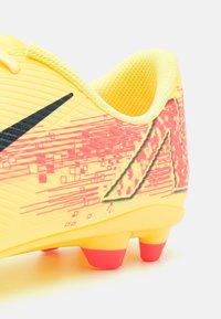 Żółte sportowe buty piłkarskie z czarnym logo, teksturowana cholewka i różowe abstrakcyjne wzory. Wyposażone w różową podeszwę z kolcami dla lepszej przyczepności.