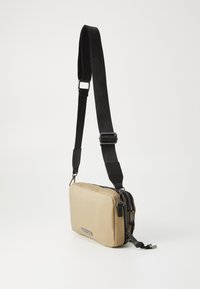 Borsa a tracolla in nylon beige con forma rettangolare, tracolla nera regolabile, doppie cerniere e una placca logo in argento sul davanti.