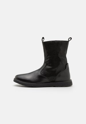 Botte noire en cuir mi-mollet avec dessus texturé, bout arrondi, semelle plate et languettes de traction de chaque côté sur un fond clair.