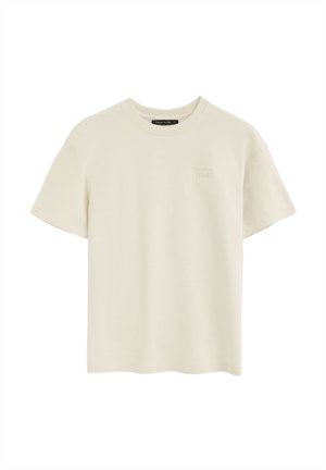 Beige T-Shirt mit kurzen Ärmeln und Rundhalsausschnitt, mit einem kleinen quadratischen Logopatch auf der linken Brust und einem schwarzen Etikett innen am Kragen.