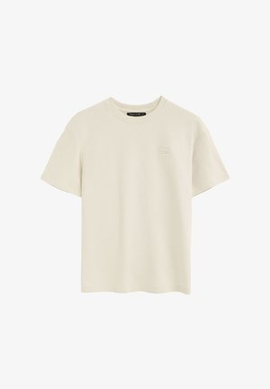 T-shirt beige à manches courtes et col rond avec un petit patch logo carré sur le côté gauche de la poitrine et une étiquette noire à l'intérieur du col.