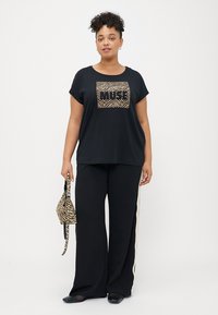 Sort T-shirt med guld zebra-mønsteret "MUSE" grafik i midten, parret med sorte brede bukser og en lille stribet håndtaske.