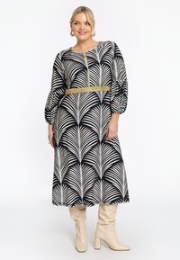 Schwarzes und weißes Kleid mit Palmblattmuster, verziert mit einem goldenen Gürtel, Glockenärmeln und knielangem Schnitt, kombiniert mit beigen Stiefeletten mit Absatz.