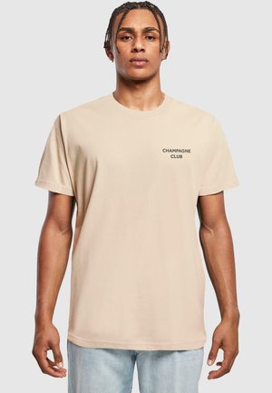 Beige bomuld t-shirt med teksten "CHAMPAGNE CLUB" i en kontrasterende skrifttype, klassisk rund hals og korte ærmer. Båret med lyseblå jeans.