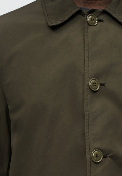 Κοντινό πλάνο πουκάμισου σε ελιά πράσινο χρώμα με κουμπιά και γιακά, εμφανίζονται τρία κουμπιά και λεπτομερής ραφή σε ομαλό ύφασμα.