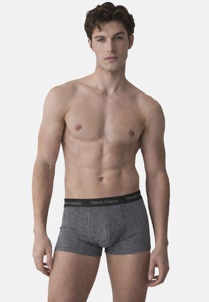 Jeune homme debout, portant un boxer gris avec une ceinture « Vincent Creation », sur fond clair uni, regardant devant lui.
