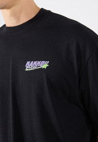 Sort bomulds T-shirt med et lilla og grønt grafisk logo på venstre bryst. Klassisk crew neck design, glat tekstur.