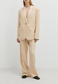 Beige gestreept pak gemaakt van lichte stof, met een blazer met één knoop, twee voorzakken en een broek met wijdere pijpen.