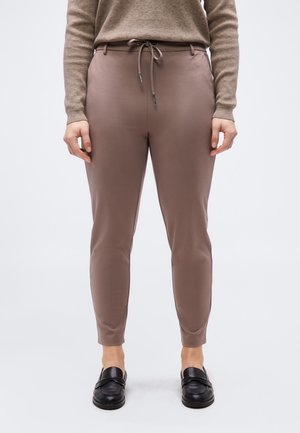 Άτομο που φορά καφέ παντελόνια με στενή εφαρμογή και κορδόνι στη μέση, ένα καφέ πουλόβερ με μακριά μανίκια που ταιριάζει και μαύρα παπούτσια χωρίς κορδόνια.