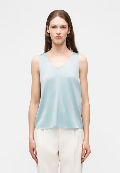 Femme portant un chemisier en satin bleu clair sans manches et un pantalon blanc, debout devant un fond blanc uni, face à l'avant.