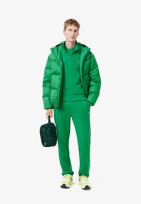 Grøn pufferjakke, matchende træningsdragt og limefarvede sneakers. Funktioner inkluderer lynlåsdetaljer og en taske med grafisk design. Glatte stofteksturer.